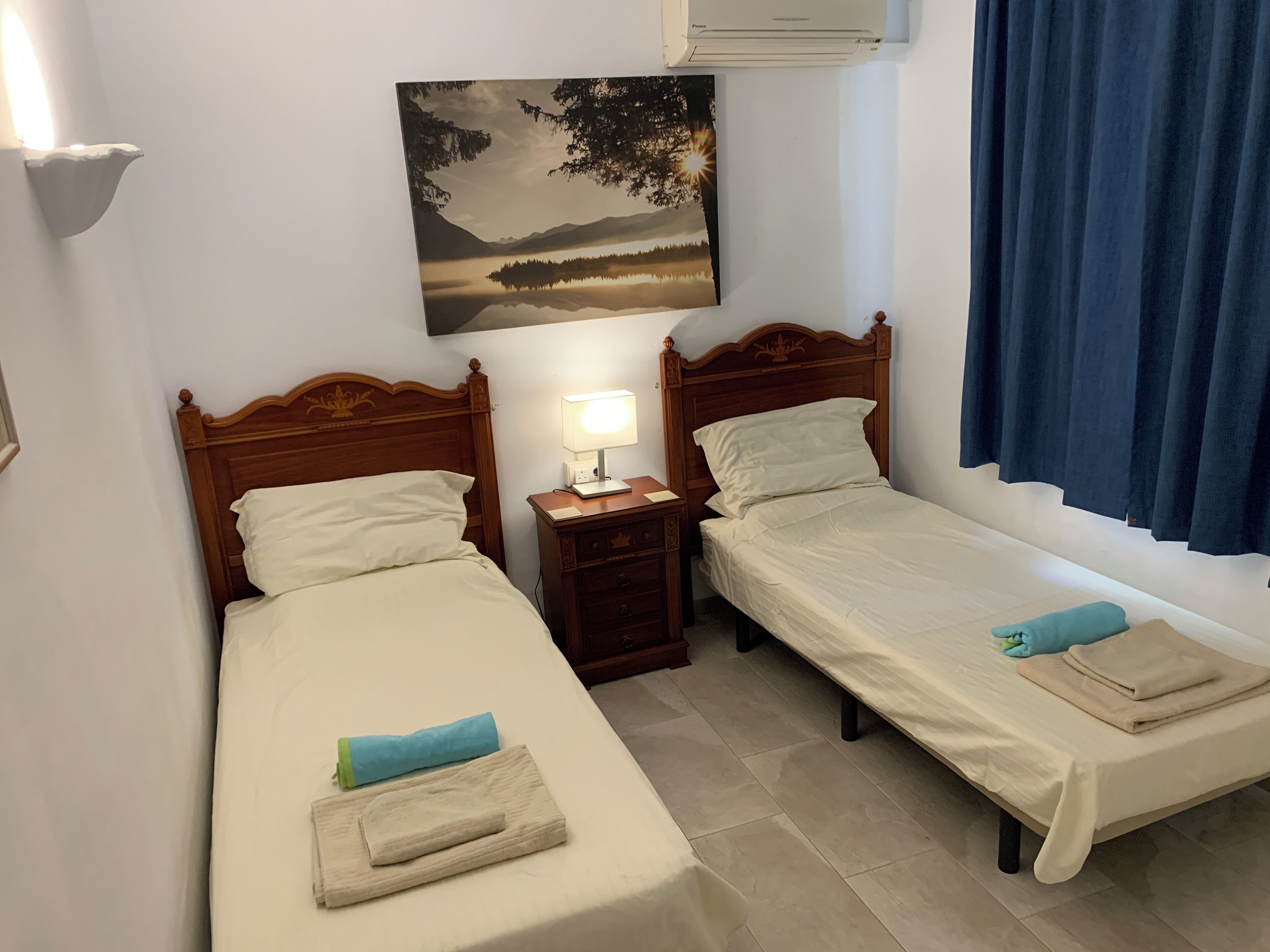 Two single beds with wooden headboards, a nightstand with a lamp, a painting of a landscape, and a window with blue curtains.
