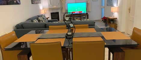 A modern living room with a dining area, a fireplace, and a television displaying a sports event.