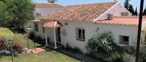 Un bâtiment blanc avec un toit en tuiles, un porche abrité et un petit jardin agrémenté de fleurs rouges.