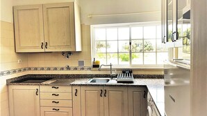 A kitchen with beige cabinets, a granite countertop, a stainless steel refrigerator, and a window with a view of greenery. - Villa located in the last condo before arriving the beach of Meco (Sesimbta)