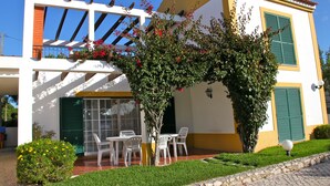 A two-story building with a white facade, green shutters, and a pergola with red flowers. There are white chairs and a table outside. - Villa located in the last condo before arriving the beach of Meco (Sesimbta)