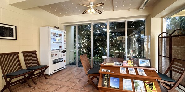 A room with a ceiling fan, a vending machine, a table with books, and two wooden chairs. - Pension Sunset Beach (Nakijin)