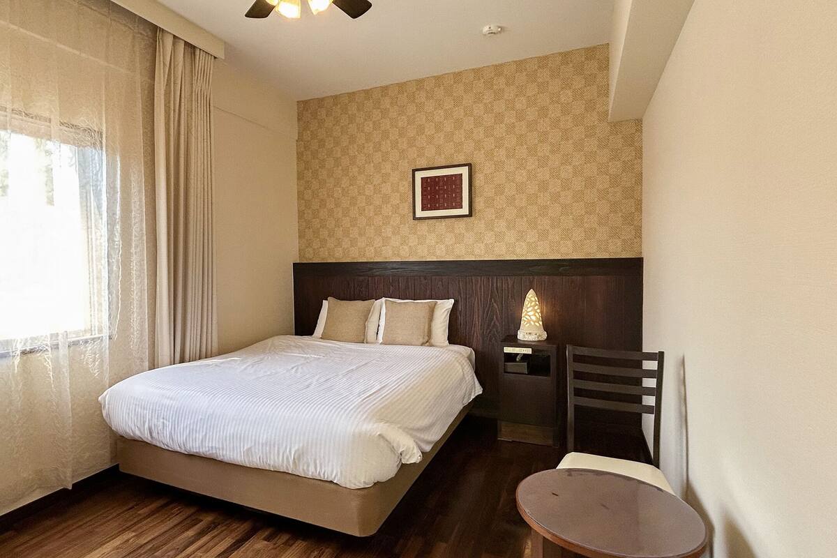Una habitación de hotel con cama, mesita de noche, una silla, una pequeña mesa y una lámpara fijada a la pared.