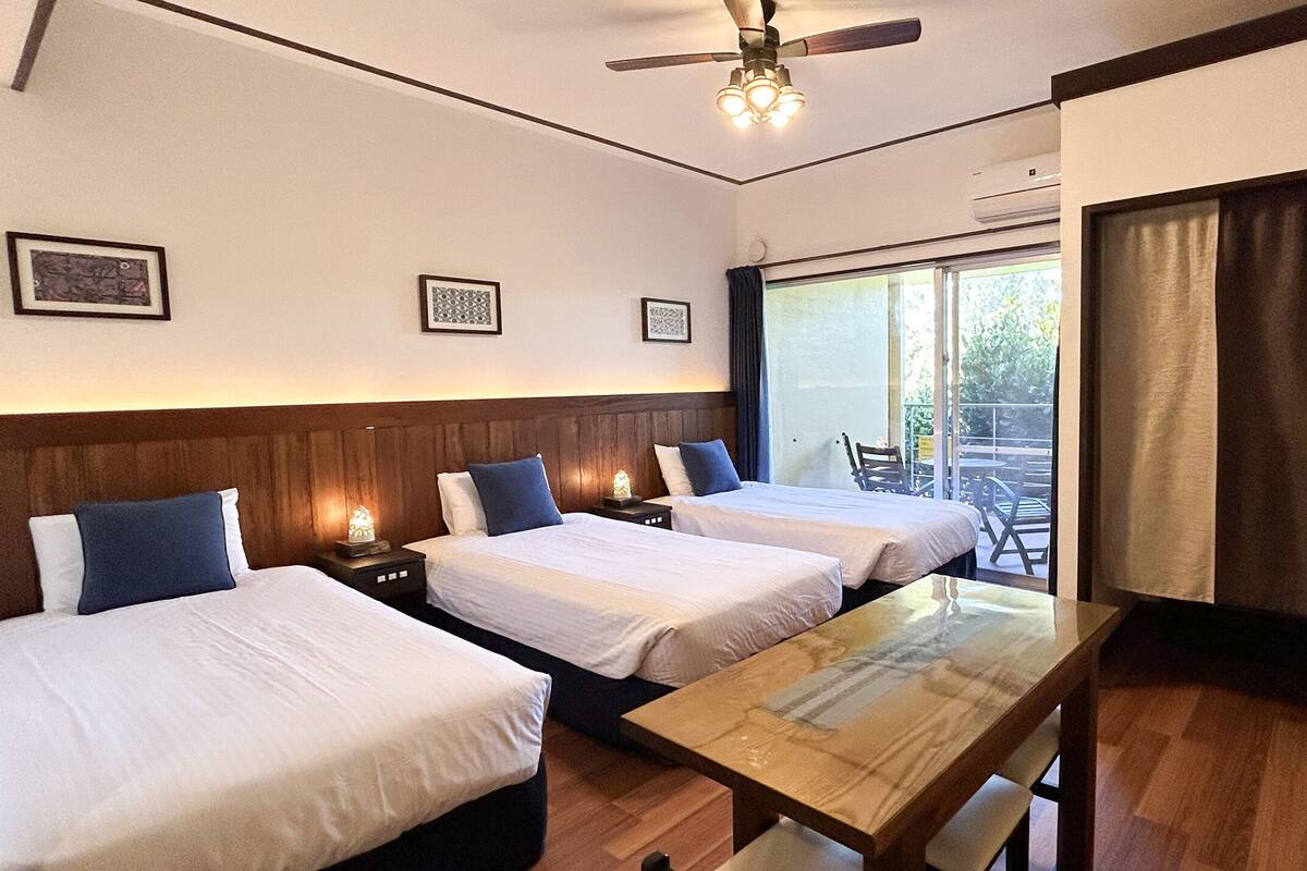 Habitación de hotel con dos camas, una mesa de madera, ventilador de techo y vistas al exterior.