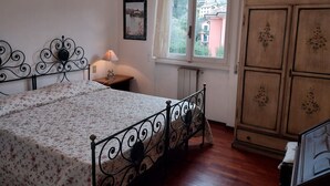 Une chambre à coucher comprenant un lit, une table de chevet, une lampe, une fenêtre donnant sur des immeubles et une armoire en bois.