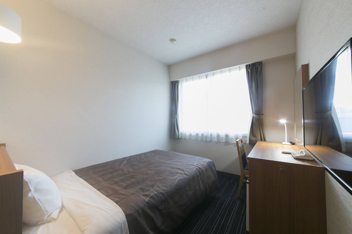 A hotel room with a bed, a desk with a lamp, a chair, and a large window with curtains.