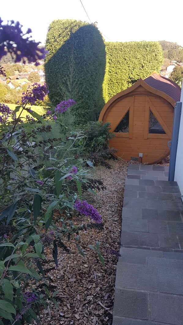 A small wooden structure with a triangular roof, surrounded by a garden with purple flowers and a stone pathway. - Apartment "Glonntal" between city, lakes & mountains (Glonn)