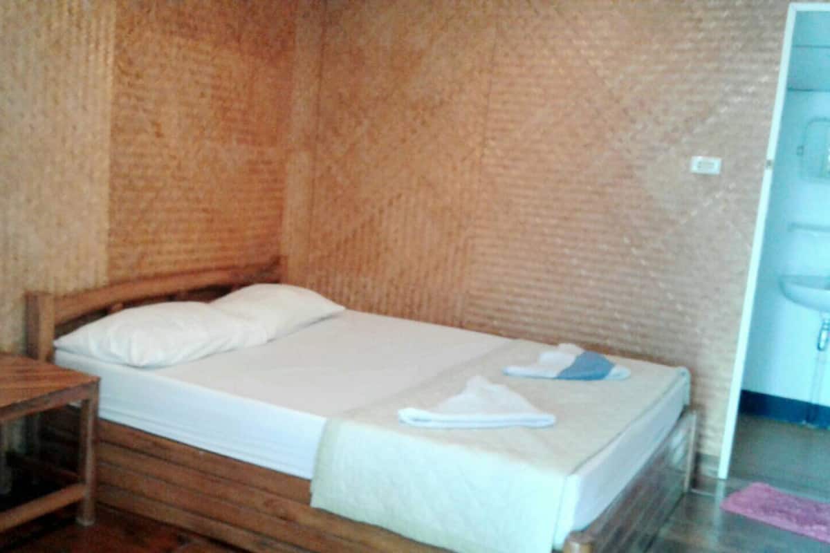 A single bed with a wooden frame, a white mattress, and a matching pillow. There is a wooden nightstand beside the bed with a bottle of water and a tissue box. A towel is placed on the bed.