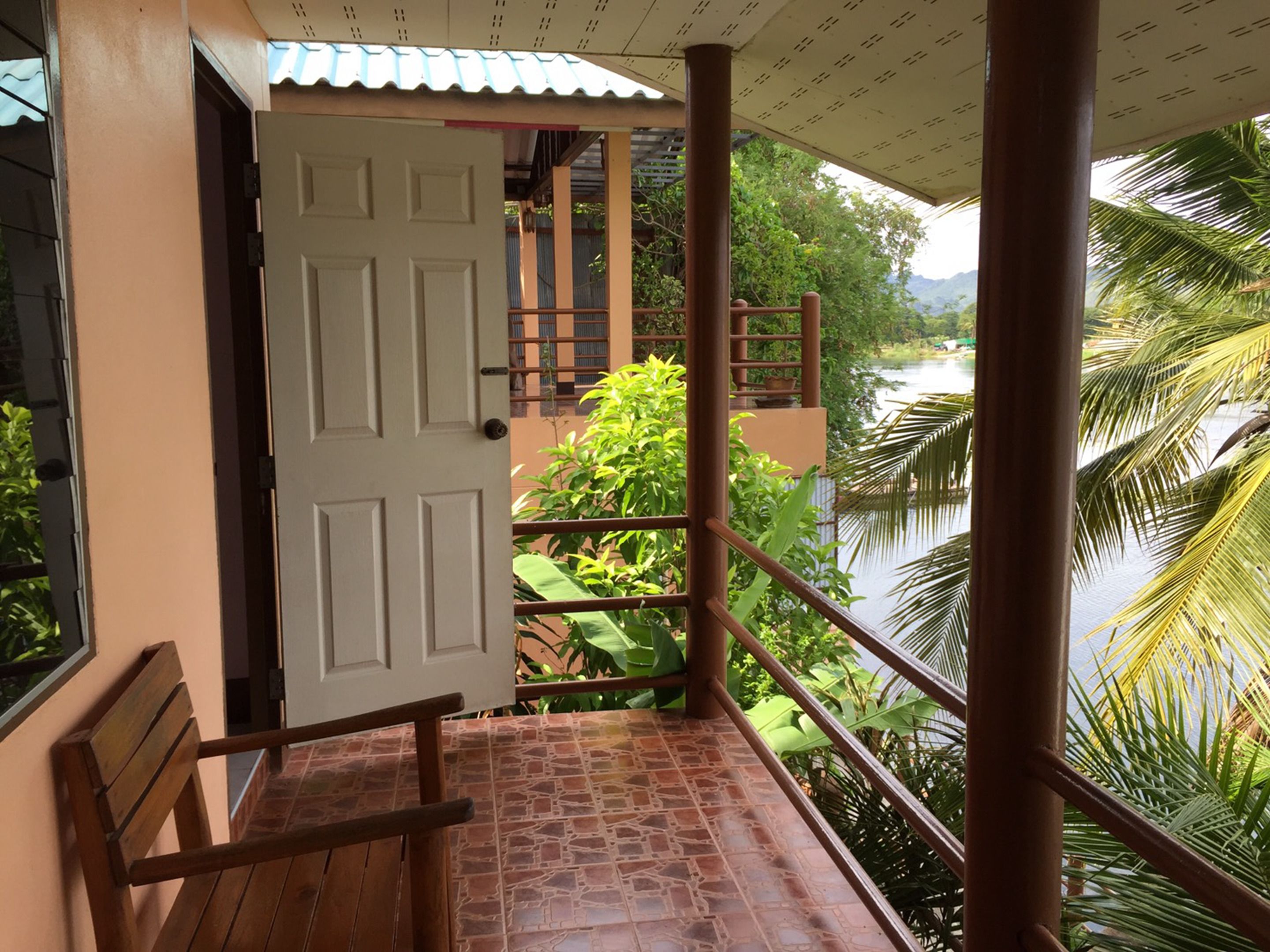 A balcony with a wooden bench, a white door, and a view of a river and greenery.