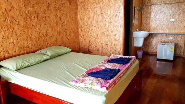 A room with a wooden wall, a bed with green bedding, a bedside table, a white sink, and a small refrigerator. - Tamarind Guest House (Kanchanaburi)