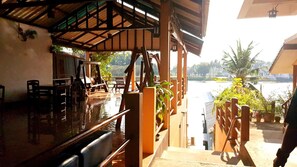 A covered outdoor seating area with wooden furniture and a view of a body of water. - Tamarind Guest House (Kanchanaburi)