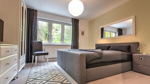 A modern bedroom with a bed, bedside tables, a chair, a dresser, and a large window with curtains.