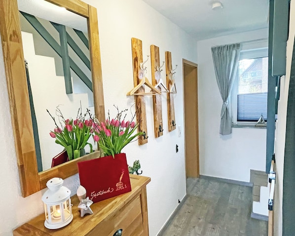 A hallway with a wooden dresser, a mirror, a vase with pink flowers, a red card, a white lantern, and a small raccoon figurine.
