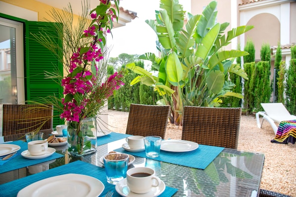 A dining table set with plates, cups, and a vase of flowers, surrounded by wicker chairs and a garden view.