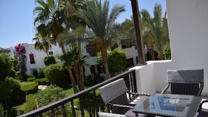 En balkon med et glasbord og to stole, med udsigt over en have med palmer og en hvid bygning i baggrunden.