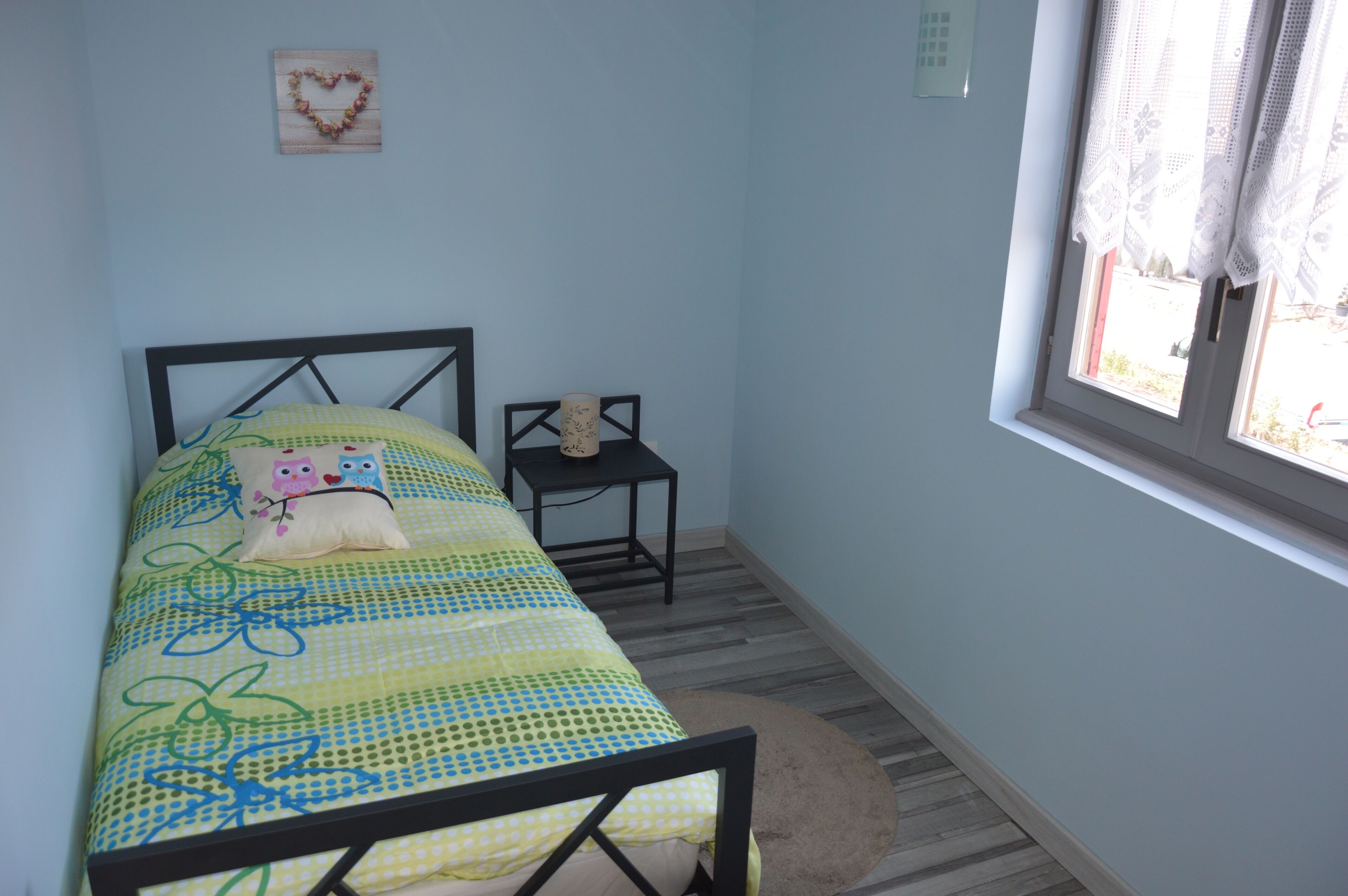 A small bedroom with a bed, a nightstand, a window, and a wall decoration.
