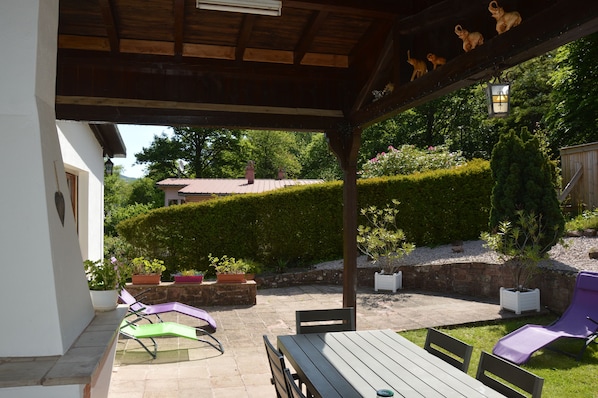 Un patio couvert avec une table et des chaises en bois, entouré de plantes en pot et d’une haie bien entretenue.