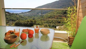 A table with a glass bowl, a glass of orange juice, a banana, an orange, and a fork on a balcony with a view of a mountainous landscape.