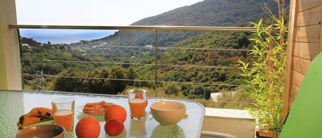 Une table avec un saladier en verre, un verre de jus d’orange, une banane, une orange et une fourchette, sur un balcon offrant une vue sur un paysage montagneux.