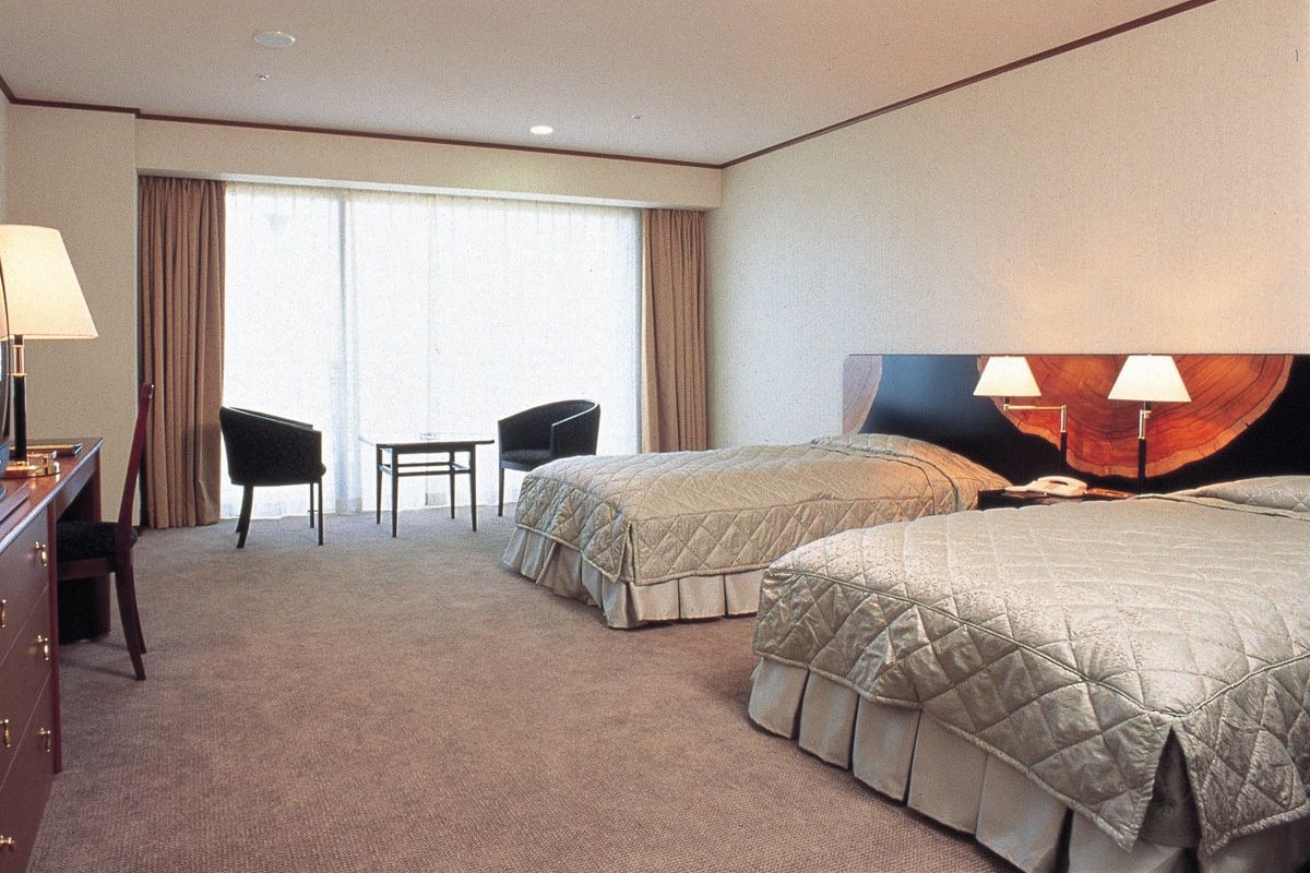 Habitación de hotel con dos camas, un escritorio, una silla y un ventanal con cortinas.