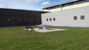 Une cour avec une table de ping-pong et un banc, entourée d’un bâtiment dont le revêtement extérieur est horizontal et dont les fenêtres sont dotées de cadres rouges.