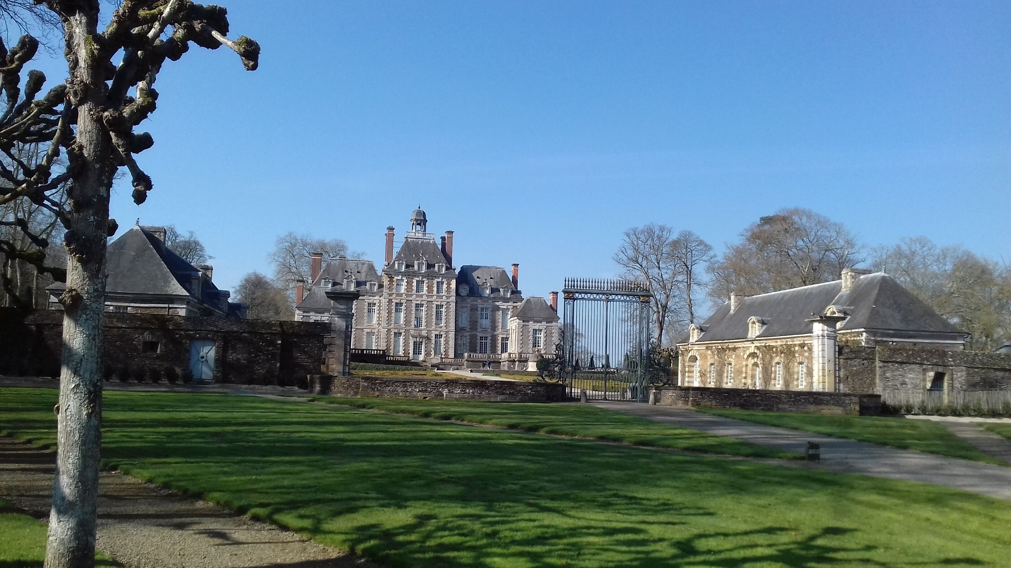 Um edifício grande e histórico com uma torre imponente e várias chaminés, cercado por jardins bem cuidados e uma entrada com portão.