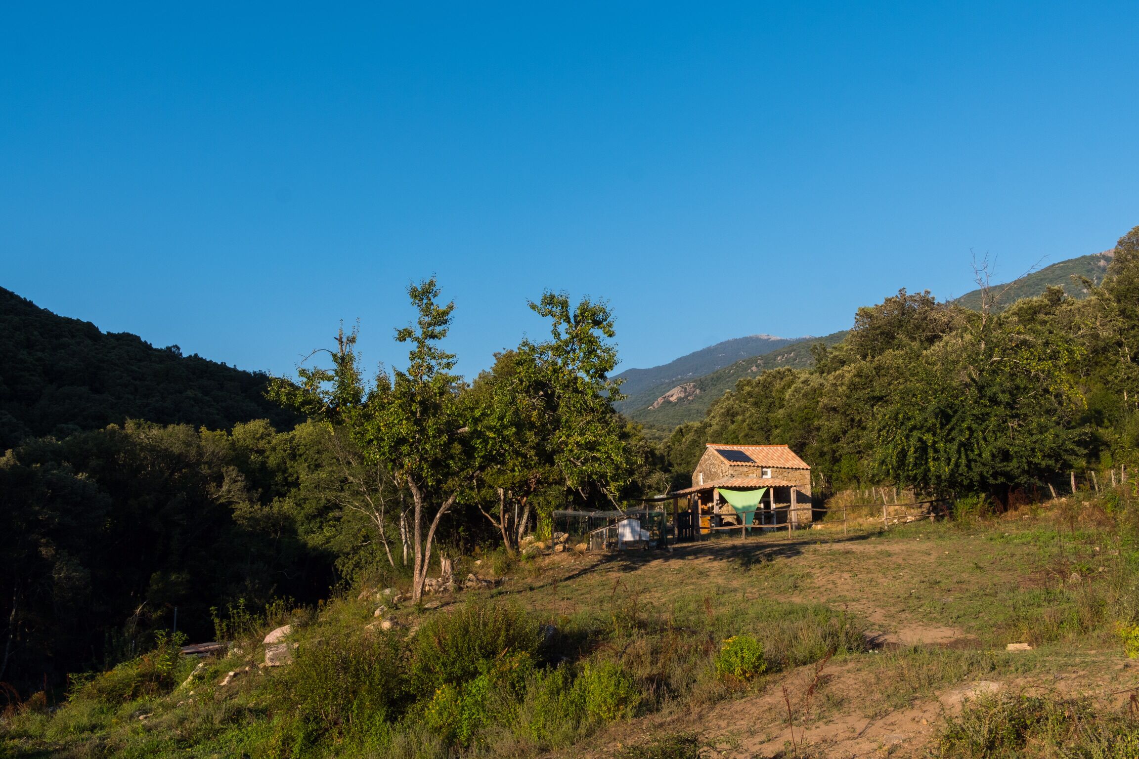 Une maison de campagne avec un toit en pente, entourée d’arbres et de collines.