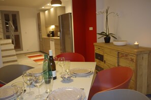Een eetruimte met een tafel voor vier personen, compleet met wijnglazen, borden en bestek. In de achtergrond staan een koelkast en een potplant.