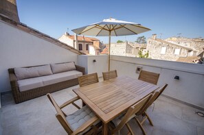Een terras op het dak, met een houten tafel en stoelen, een rieten bank met kussens en een grote parasol.