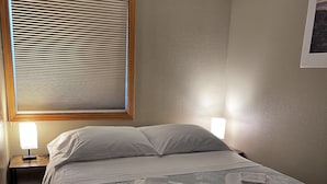 A neatly made bed with a blue patterned bedspread and two folded towels on the foot of the bed. There is a nightstand with a lamp and a window with blinds. - 1 Block from the Bay!  (Seward)