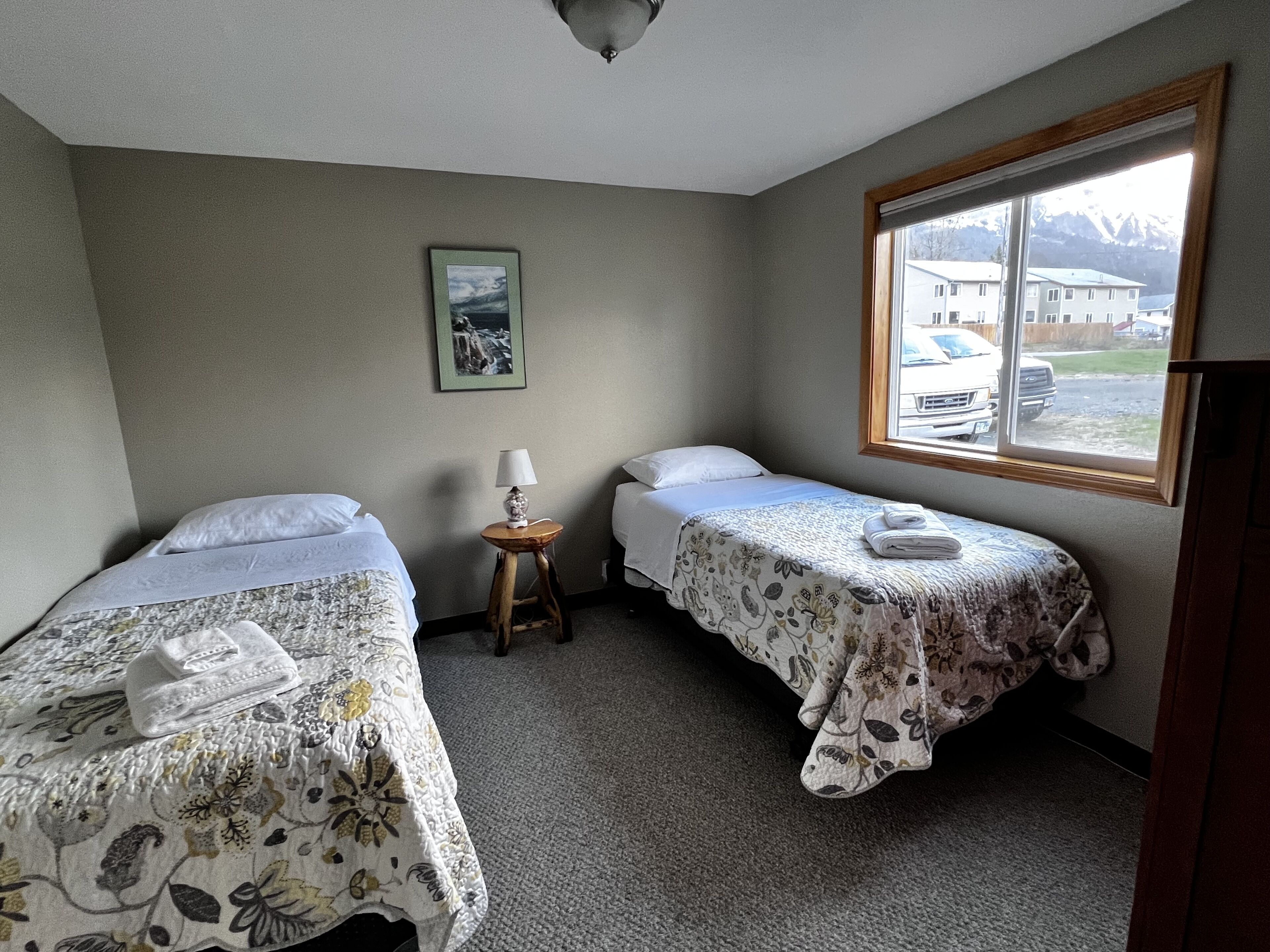 A hotel room with two single beds, a small table with a lamp, a framed picture on the wall, and a window with a view of buildings and mountains.