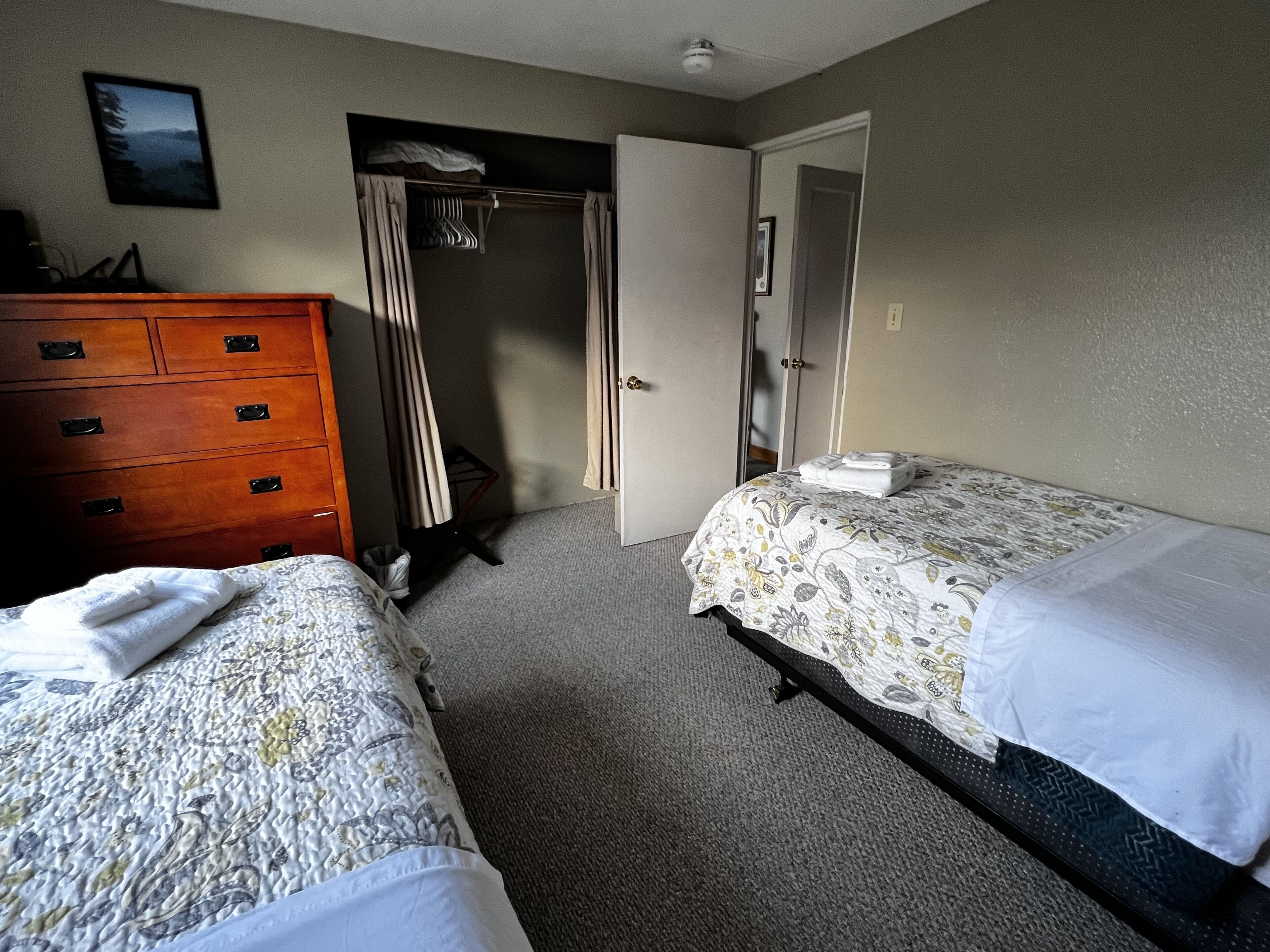 A hotel room with two beds, a wooden dresser, a closet, and a door leading to another room.