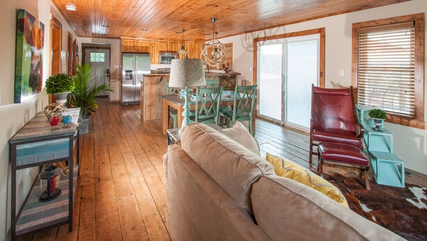 A spacious living area with a wooden ceiling, a sofa, a dining table, and a kitchen in the background.