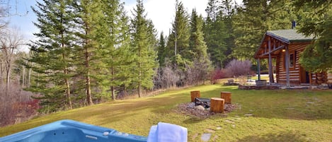 A hot tub in a forested area with a wooden cabin and a grassy lawn.