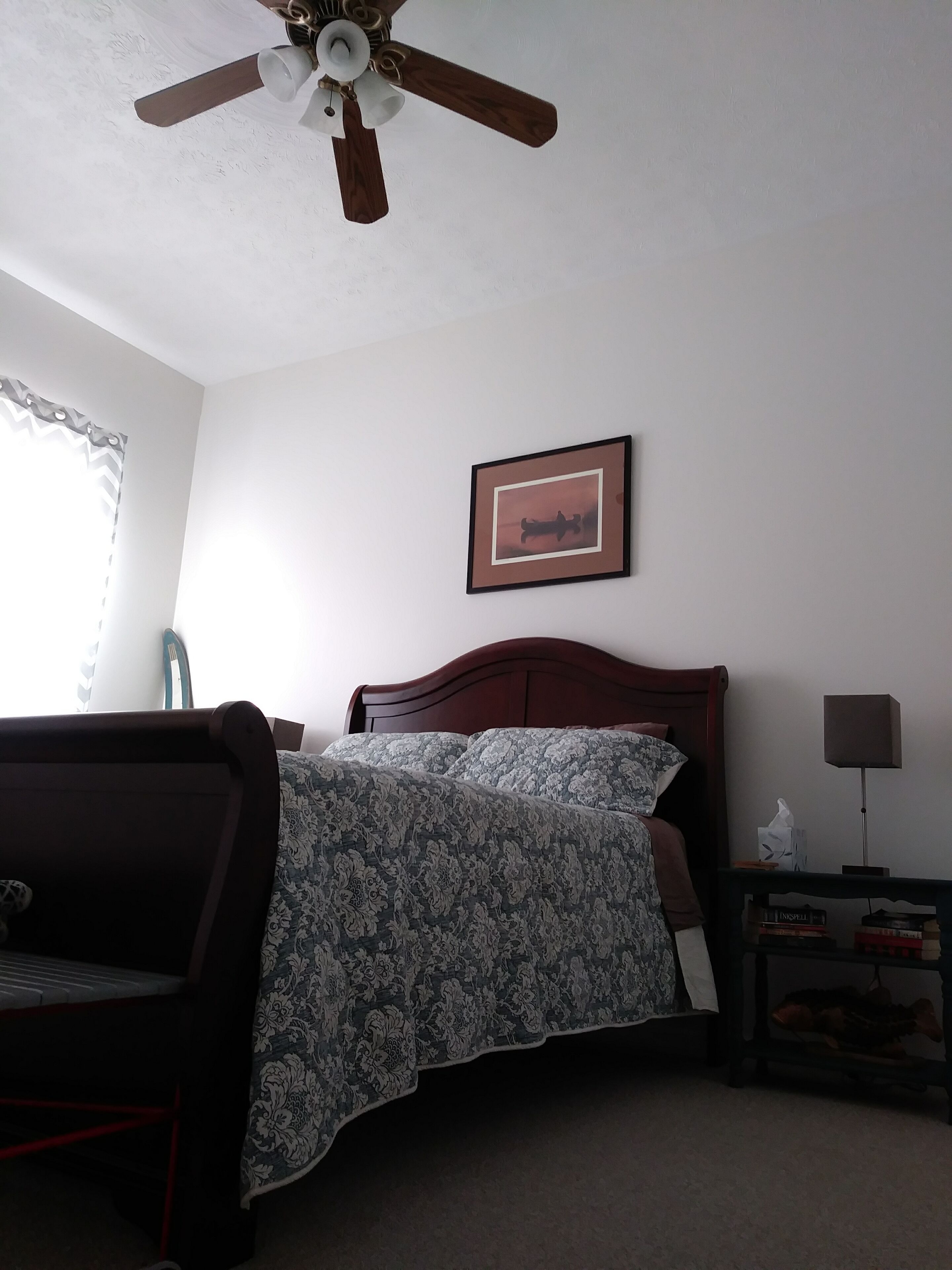 A bedroom with a bed, a chair, a nightstand, a lamp, and a ceiling fan.