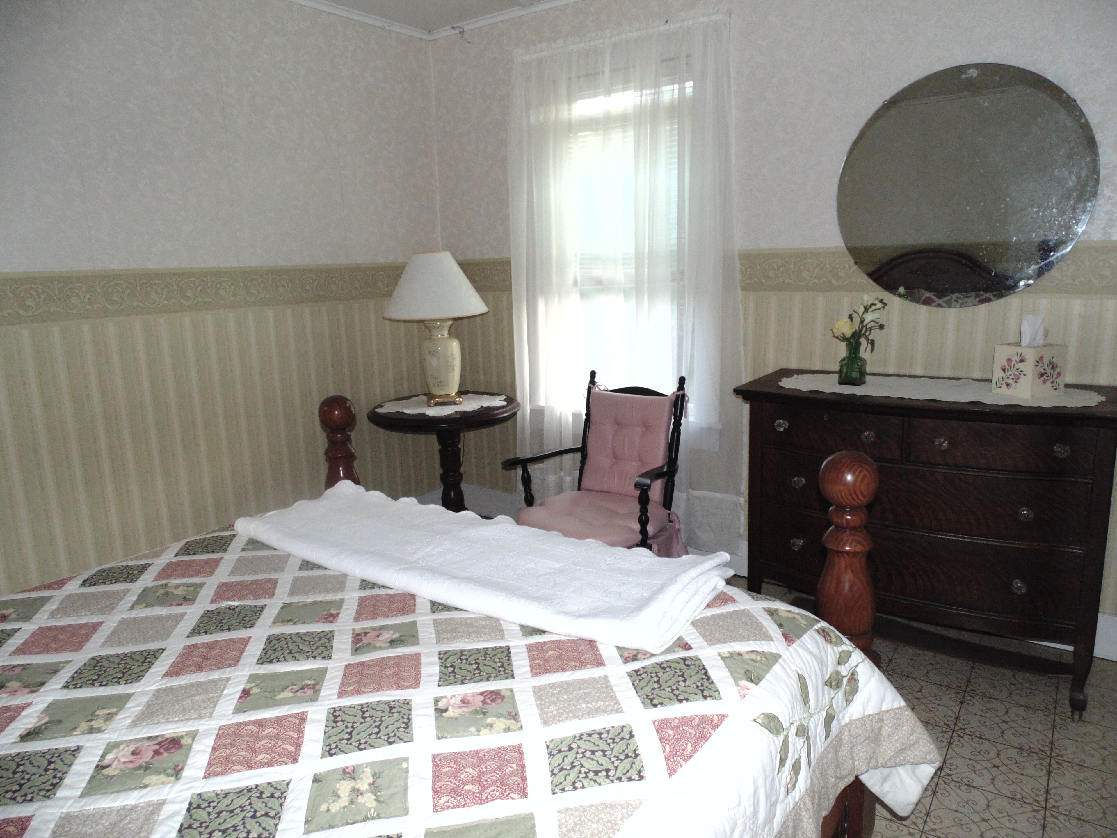 Una cama bien hecha con colcha floral, una silla de madera, una mesita de noche con lámpara, un aparador y un espejo.