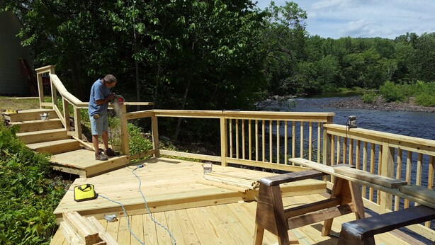 Une terrasse en bois avec une rambarde, une personne utilisant un outil électrique, et une rivière en arrière-plan.