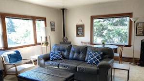 A living room with a leather sofa, ottoman, and a dining table with chairs. There are windows with views of greenery outside.
