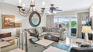 Een ruime woonkamer met een grote hoekbank, een glazen salontafel en een kroonluchter. Er is een plafondventilator en een groot raam met gordijnen.