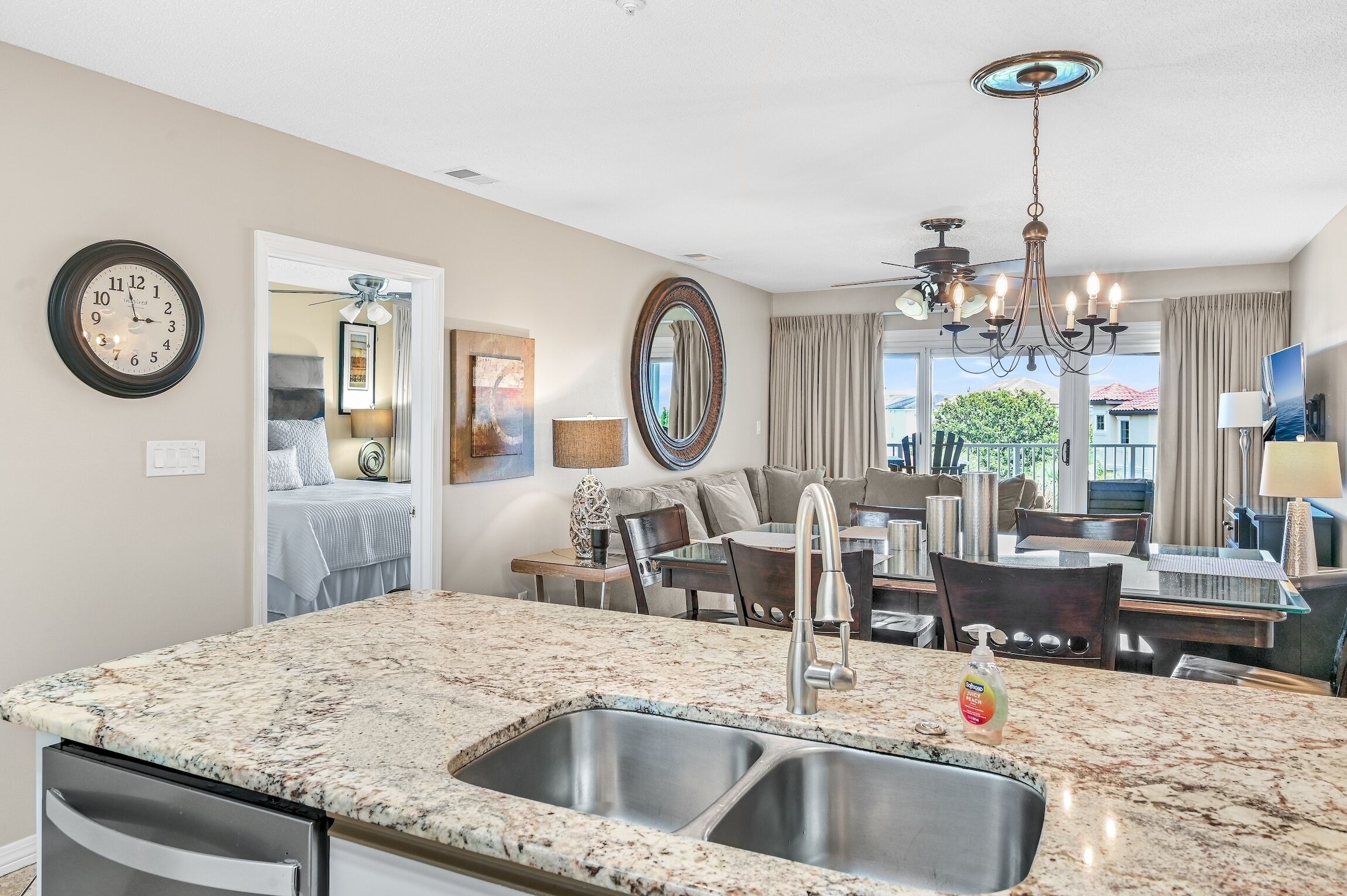 A modern kitchen with granite countertops, stainless steel sink, and a chandelier. There is a dining area with a table and chairs, and a living room with a sofa and a TV.