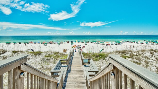 Een houten looppad dat naar een strand leidt, met parasols en mensen op het strand.