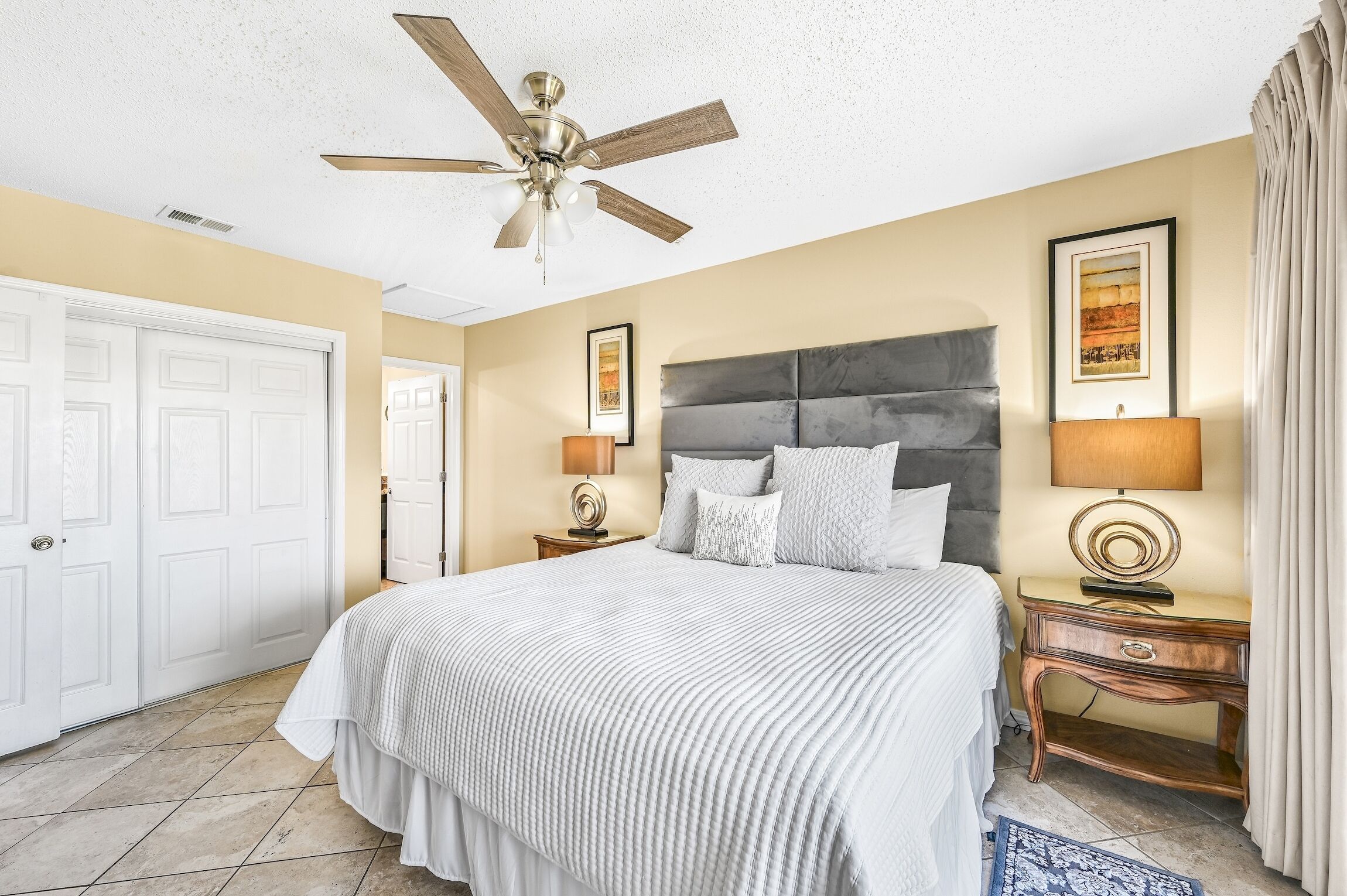 A bedroom with a bed, a ceiling fan, a nightstand with a lamp, and a door.