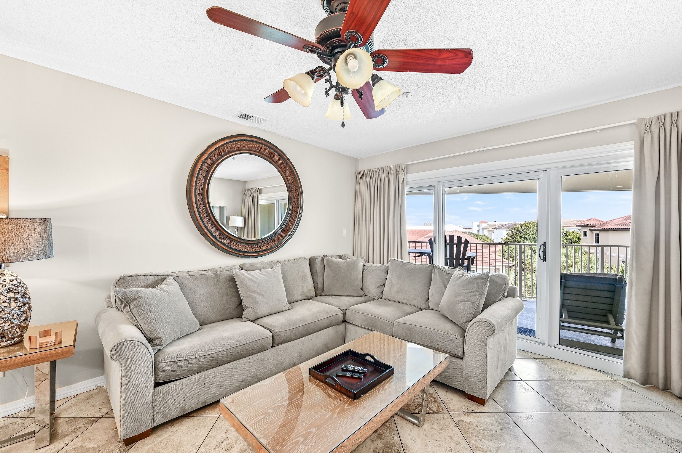 A living room with a sectional sofa, a glass coffee table, a ceiling fan, a round mirror, and a balcony with a view.