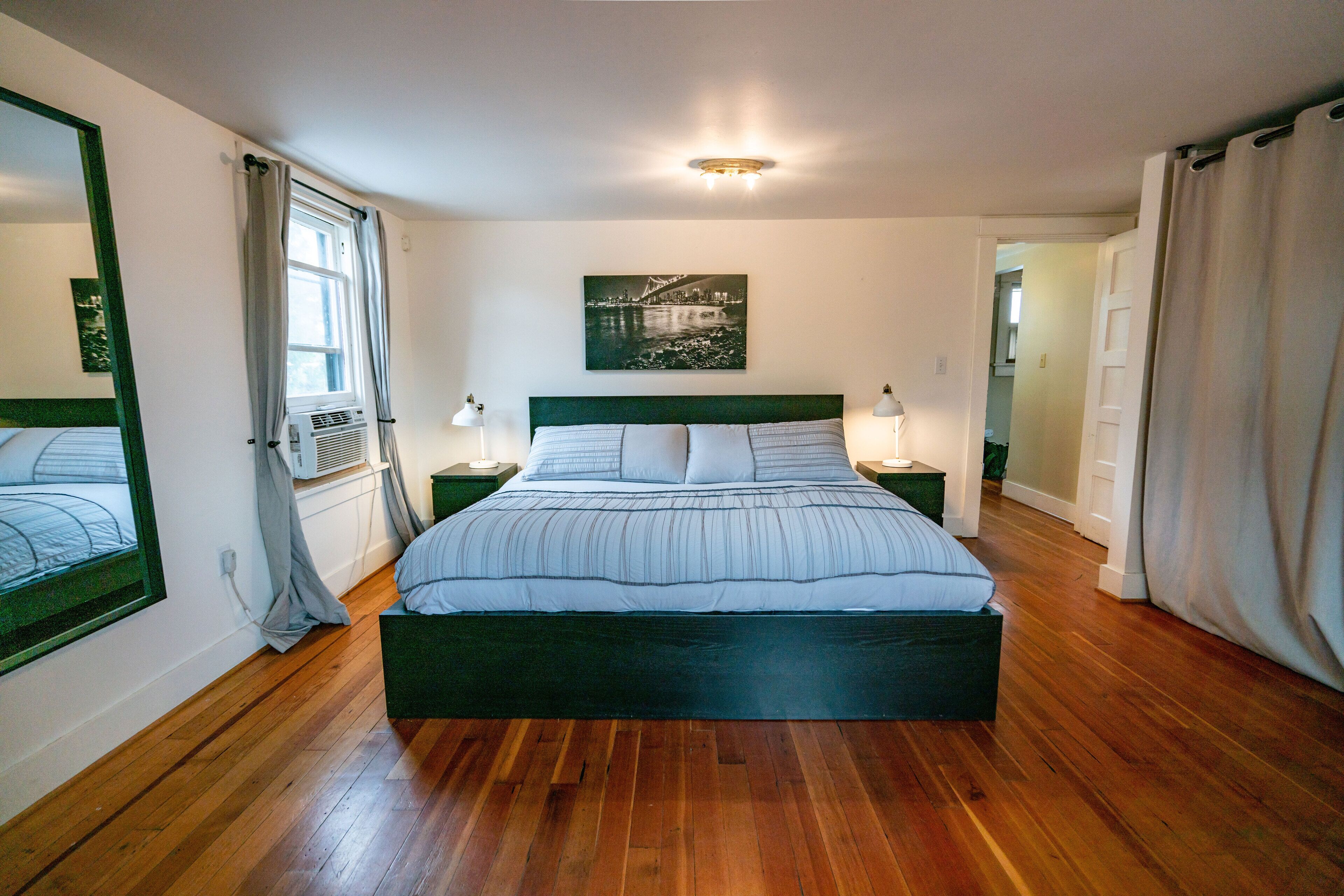 Une chambre à coucher avec un grand lit, une tête de lit, deux tables de chevet avec des lampes, une fenêtre avec des rideaux, un tableau accroché au mur et une porte donnant sur une autre pièce.