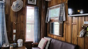 A wooden-paneled room with a sofa, a table with a kettle, a fan, and a television. - Sichang My Home (Ko Sichang)