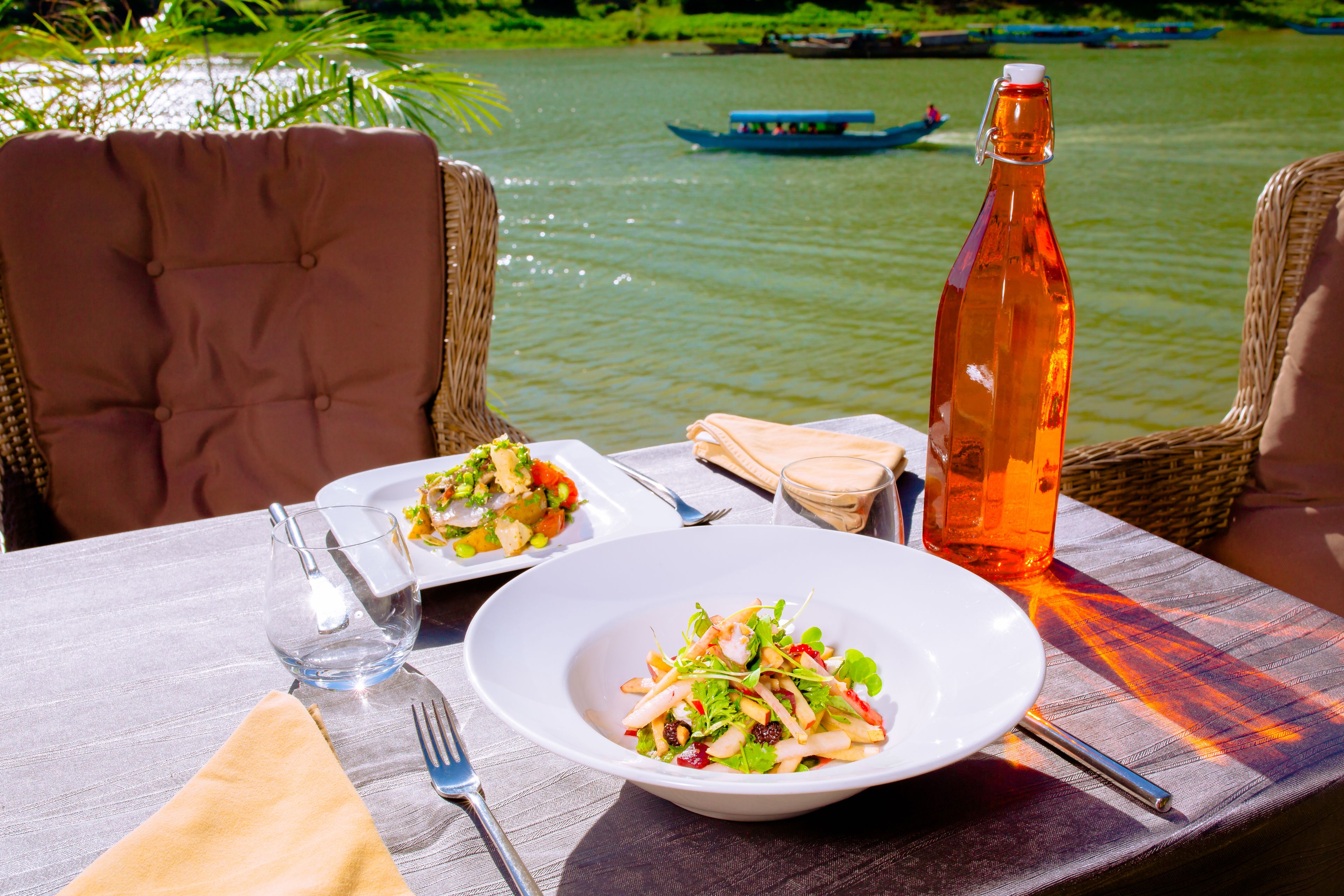 A table set with plates of food, a bottle, and cutlery, with a view of a river and boats in the background.