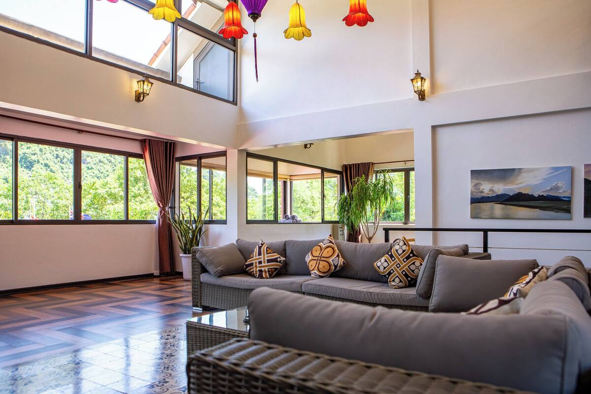 A spacious living room with a grey sofa, large windows, and a view of greenery.