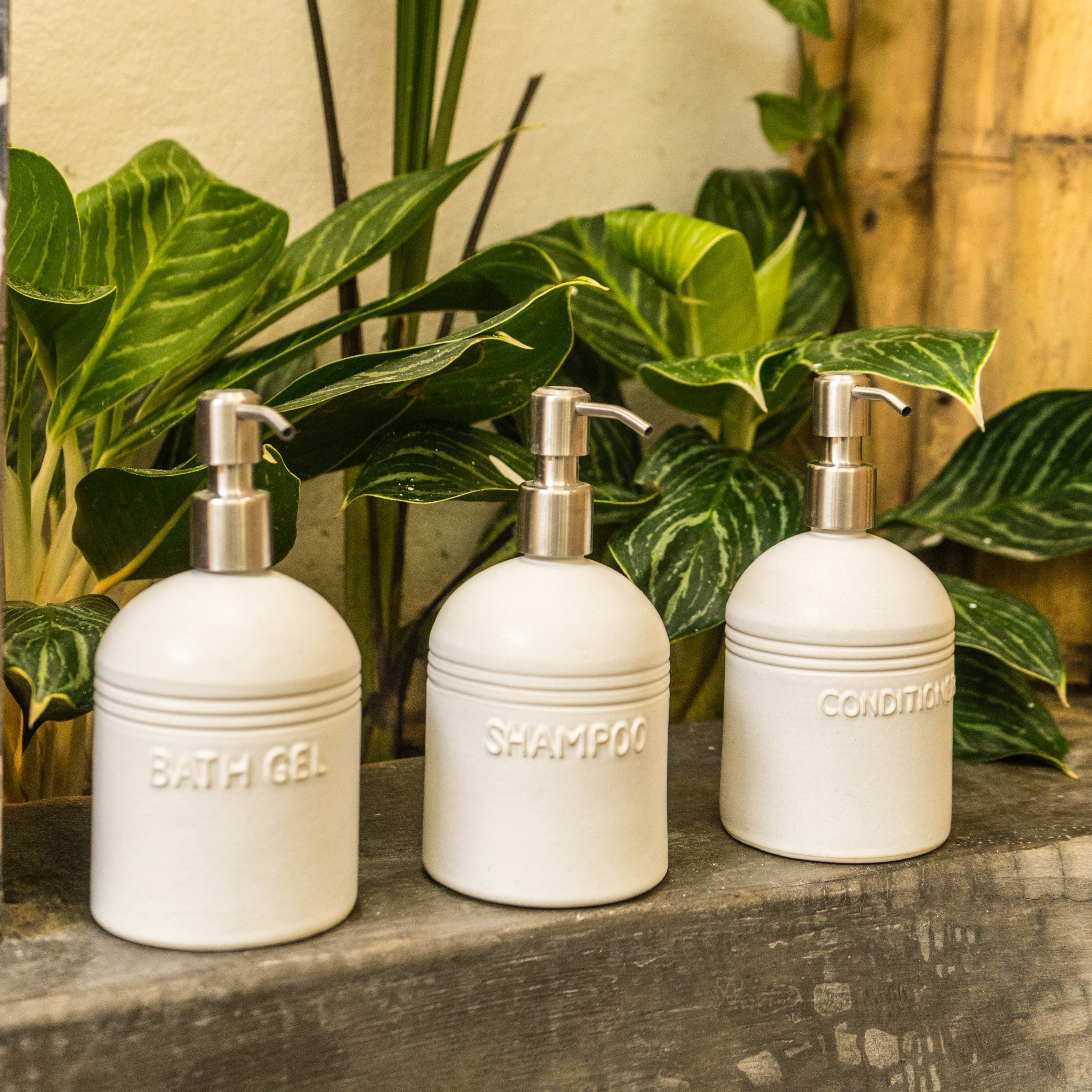 Drei weiße Flaschen mit den Aufschriften 'Bath Gel', 'Shampoo' und 'Conditioner' auf einer Holzoberfläche, im Hintergrund grüne Pflanzen.