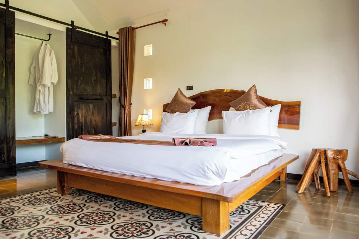 A large bed with a wooden frame and white bedding, a wooden bench at the foot of the bed, a wooden stool, and a large wooden door.