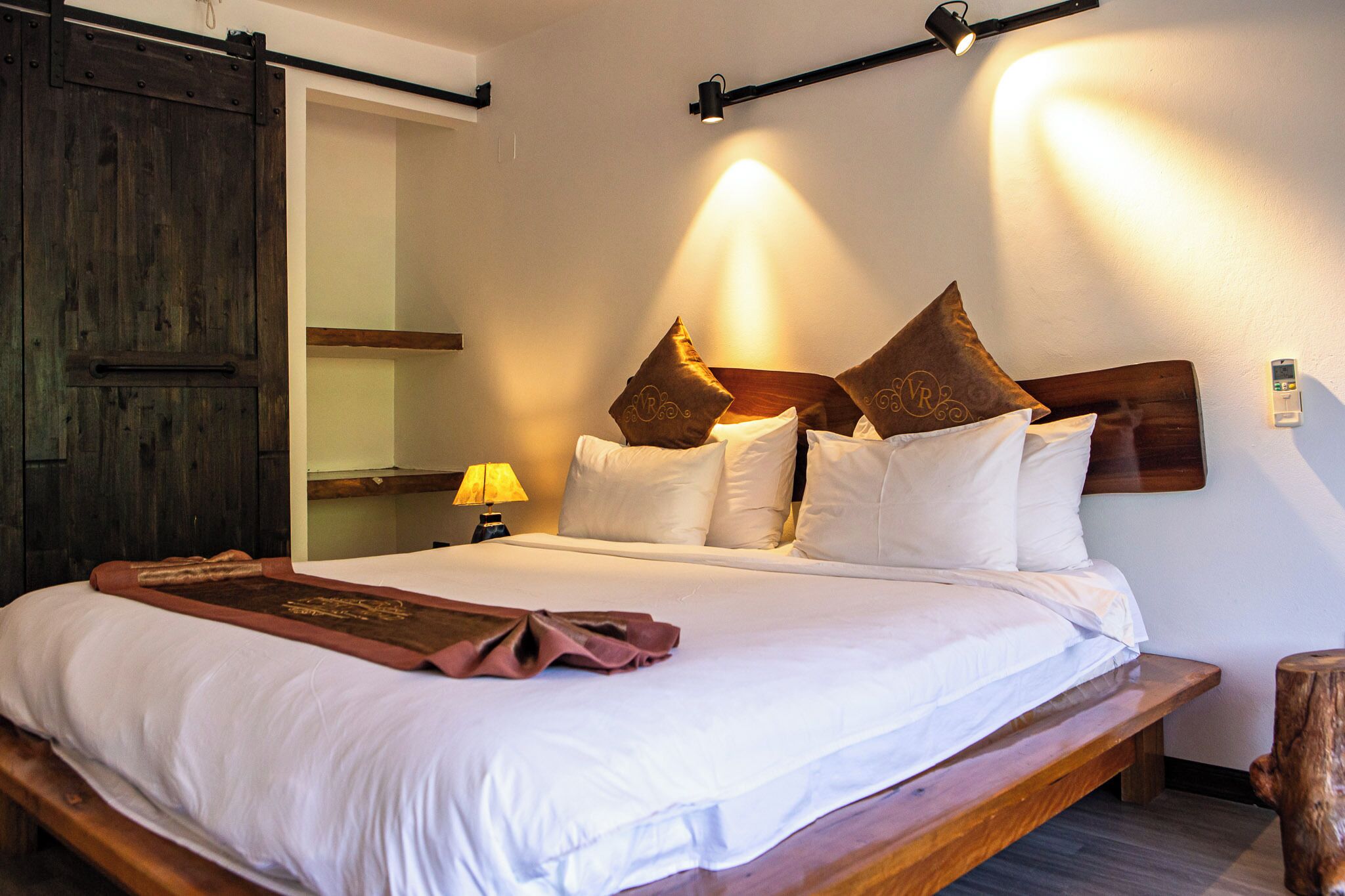 A neatly made bed with white linens and brown decorative pillows, a wooden headboard, and a bedside lamp.